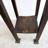 A dark brown wooden chair viewed from above, featuring a square seat with distressed gold-toned wear and four straight legs with square feet, set against a white wall and concrete floor.