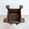 A top-down view of a dark brown wooden chair with blocky, geometric armrests and a slatted back, set against a white wall and concrete floor.