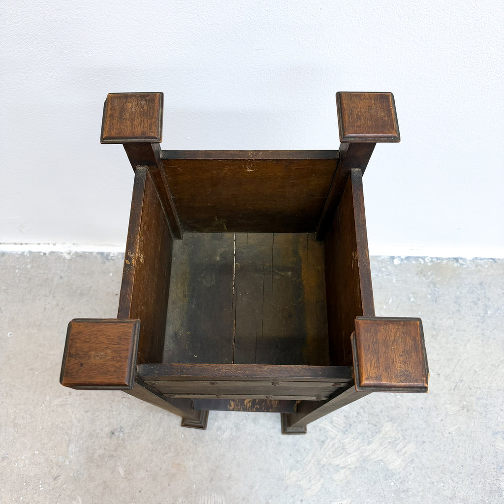A top-down view of a dark brown wooden chair with blocky, geometric armrests and a slatted back, set against a white wall and concrete floor.