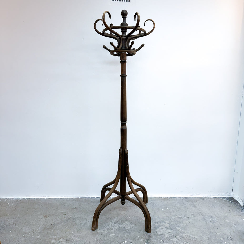 A dark brown wooden coat rack with a multi-hook top and a tripod-style base, standing on a concrete floor against a plain white wall.