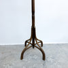 A vintage dark brown wooden coat rack with a curved tripod base, photographed against a white wall and concrete floor.