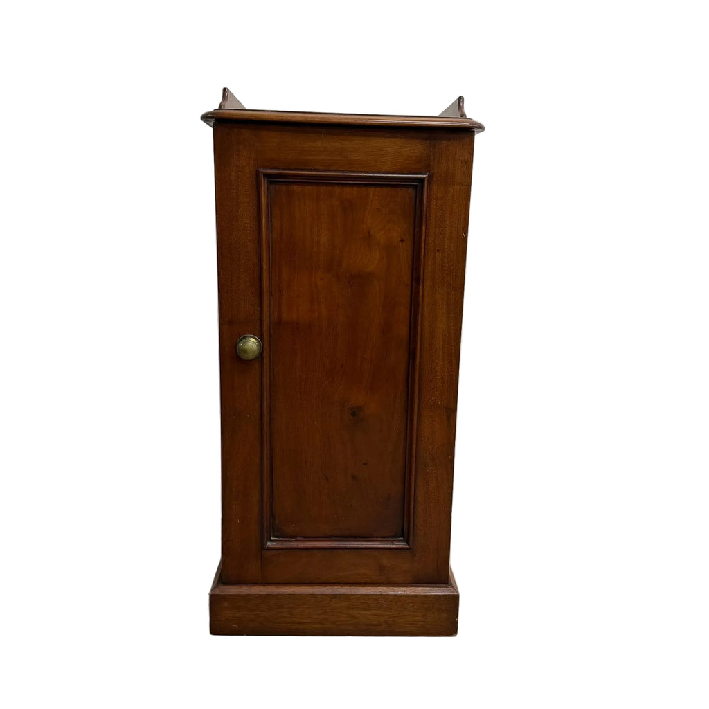 A vintage wooden cabinet with a single paneled door, turned brass knob, and decorative finials on the top, set against a plain white wall and concrete floor.