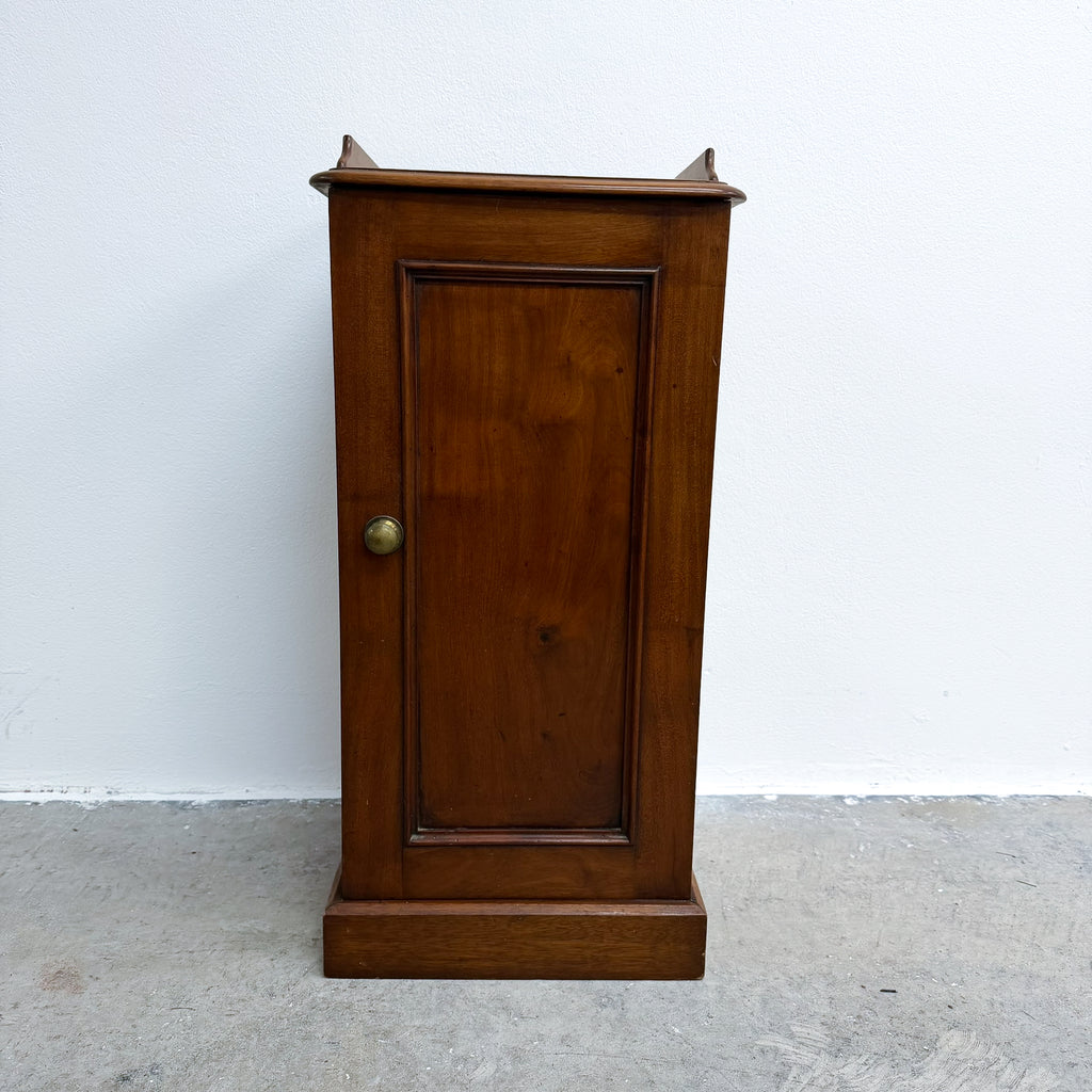 A vintage wooden cabinet with a single paneled door, turned brass knob, and decorative finials on the top, set against a plain white wall and concrete floor.
