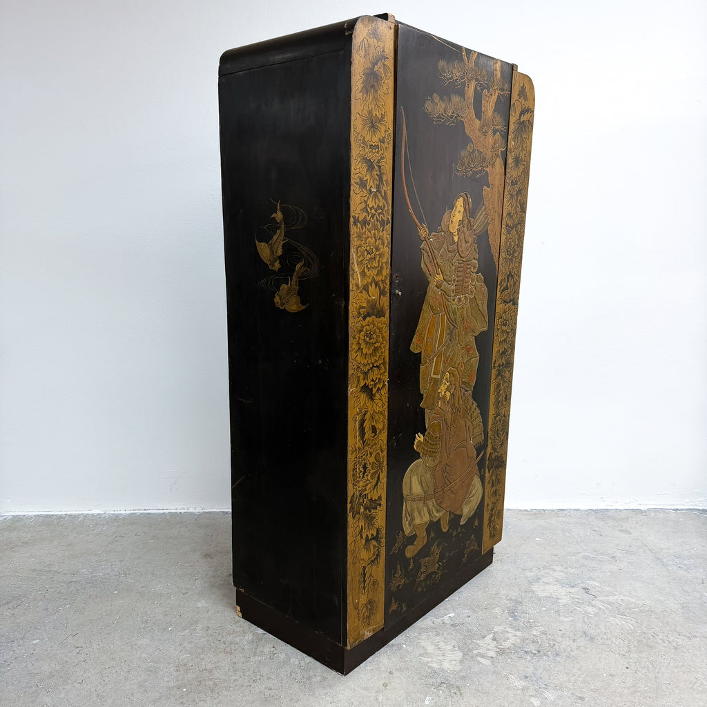 A vintage black lacquered folding cabinet with gold floral borders and hand-painted gold figures, photographed against a plain white wall and concrete floor.