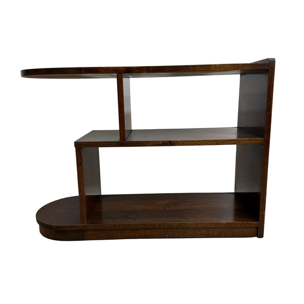 A mid-century modern wooden side table or shelf with a dark brown finish, featuring a curved top, two open shelves, and a rounded base, set against a plain off-white wall and concrete floor.