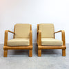Two mid-century modern wooden armchairs with beige fabric cushions, positioned side-by-side against a plain white wall and gray concrete floor.