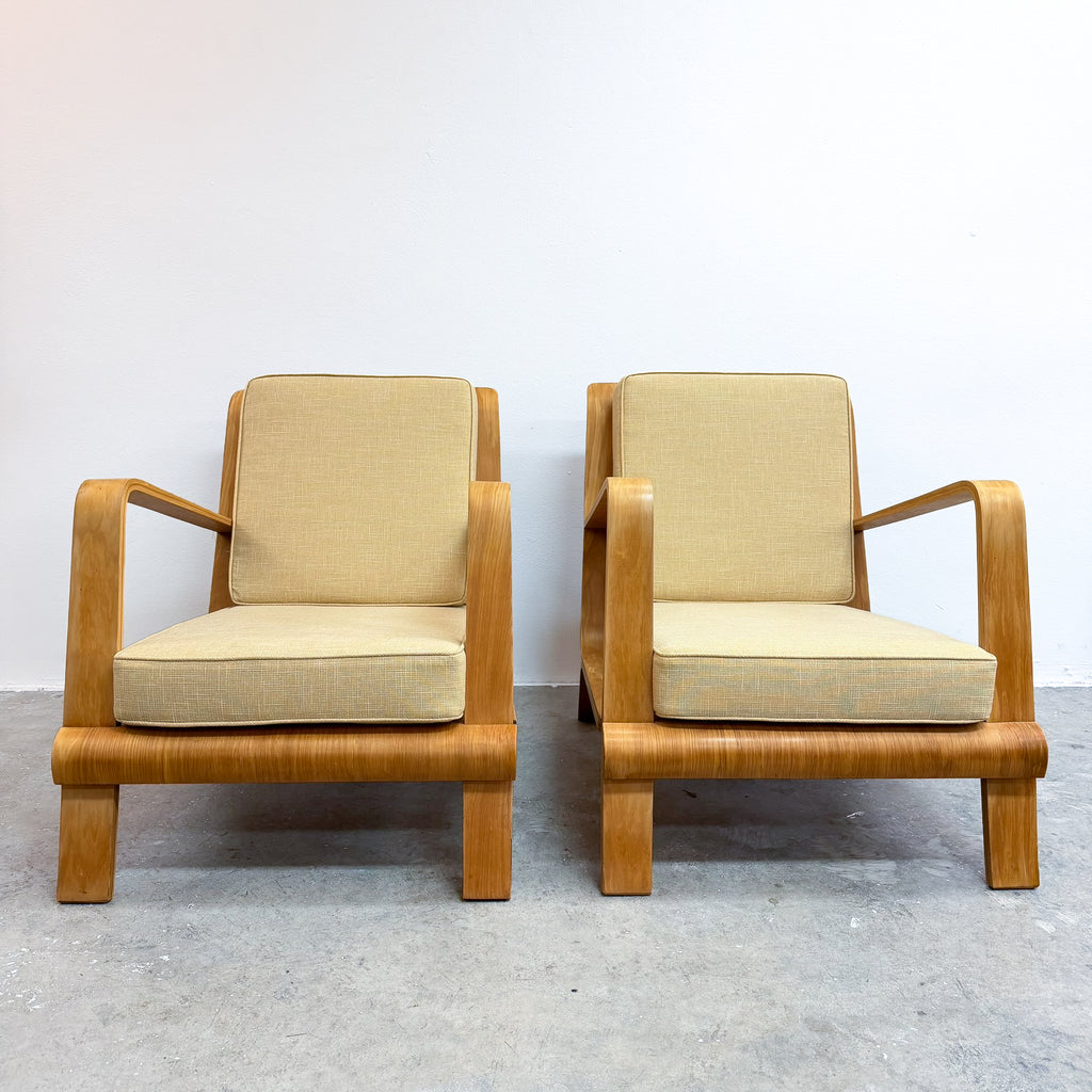 Two mid-century modern wooden armchairs with beige fabric cushions, positioned side-by-side against a plain white wall and gray concrete floor.
