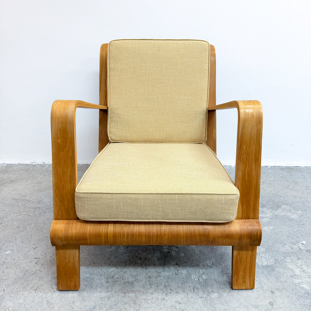 A mid-century modern lounge chair with curved wooden arms and legs, upholstered in a light beige textured fabric, set against a plain white wall and gray concrete floor.