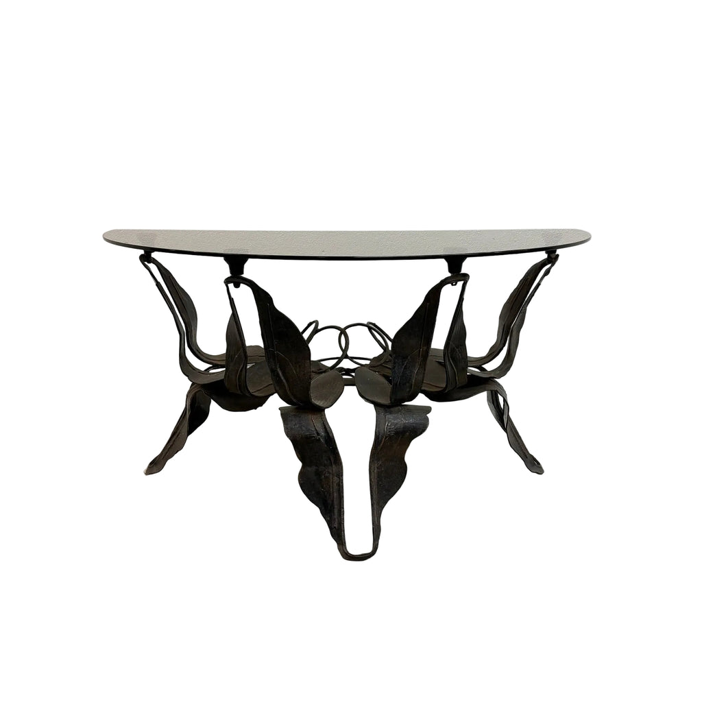 A mid-century modern metal dining table with a curved, sculptural leaf-like base and a light gray oval glass top, set against a plain white wall and concrete floor.