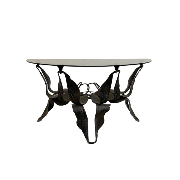 A mid-century modern metal dining table with a curved, sculptural leaf-like base and a light gray oval glass top, set against a plain white wall and concrete floor.