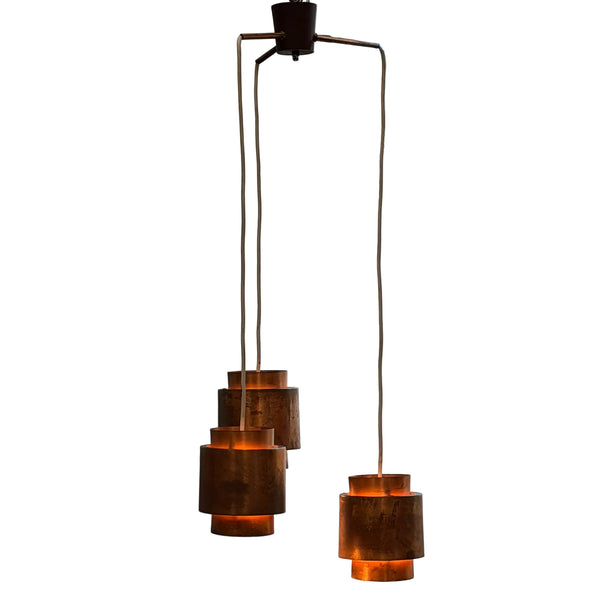 A mid-century modern multi-light pendant chandelier with three tiers of cylindrical copper shades hanging from a brass ceiling canopy against a plain white wall.