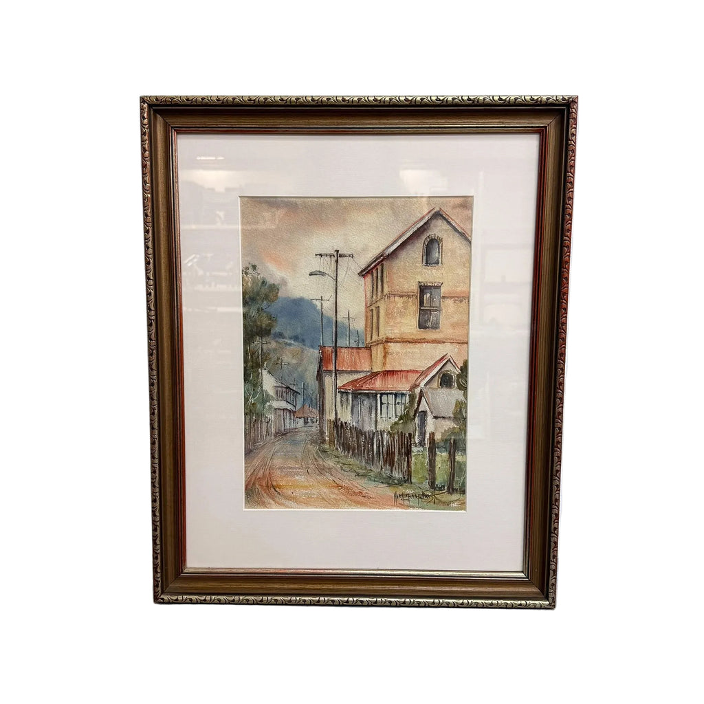 A framed watercolor painting of a quiet street scene with old houses, a dirt road, and utility poles, set in a dark brown ornate frame against a white wall.