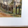 A close-up of a framed watercolor painting showing a rustic fence, house, and dirt path, with a white mat and a metallic frame edge visible.