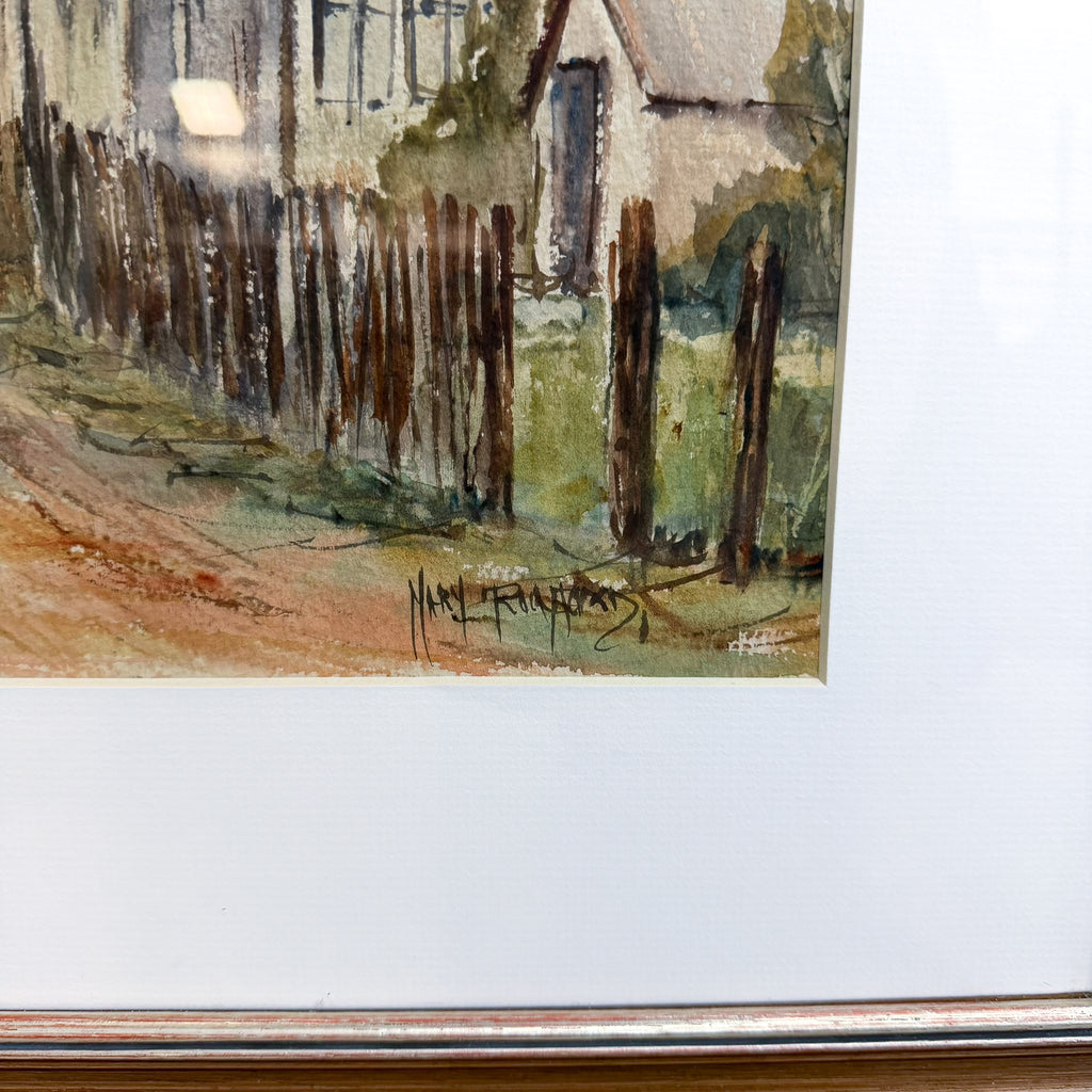 A close-up of a framed watercolor painting showing a rustic fence, house, and dirt path, with a white mat and a metallic frame edge visible.