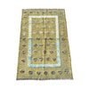 A rectangular vintage-style area rug with a distressed olive green base, featuring a central light blue border, rows of small floral motifs, and fringed edges, laid flat on a gray concrete surface.