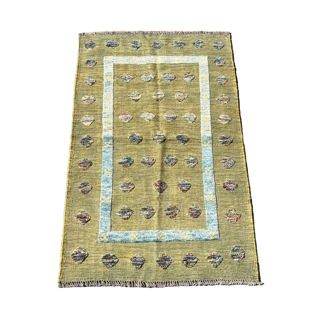 A rectangular vintage-style area rug with a distressed olive green base, featuring a central light blue border, rows of small floral motifs, and fringed edges, laid flat on a gray concrete surface.