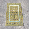 A rectangular vintage-style area rug with a distressed olive green base, featuring a central light blue border, rows of small floral motifs, and fringed edges, laid flat on a gray concrete surface.