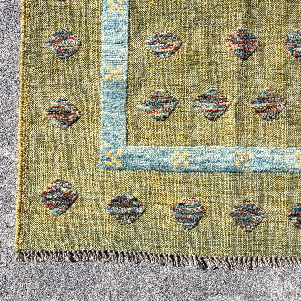 A close-up of a woven rug corner showing a mustard yellow field with a light blue border and multicolored oval motifs, photographed on a gray stone surface.