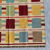A close-up of a woven kilim rug with a geometric pattern of colorful squares and stripes on a cream background, showing its texture and fringe detail.