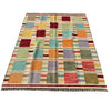 A rectangular woven area rug with a multicolor patchwork design, laid flat on a gray concrete surface. The rug features a grid of colored squares in orange, teal, red, green, purple, and beige, finished with a fringed edge.