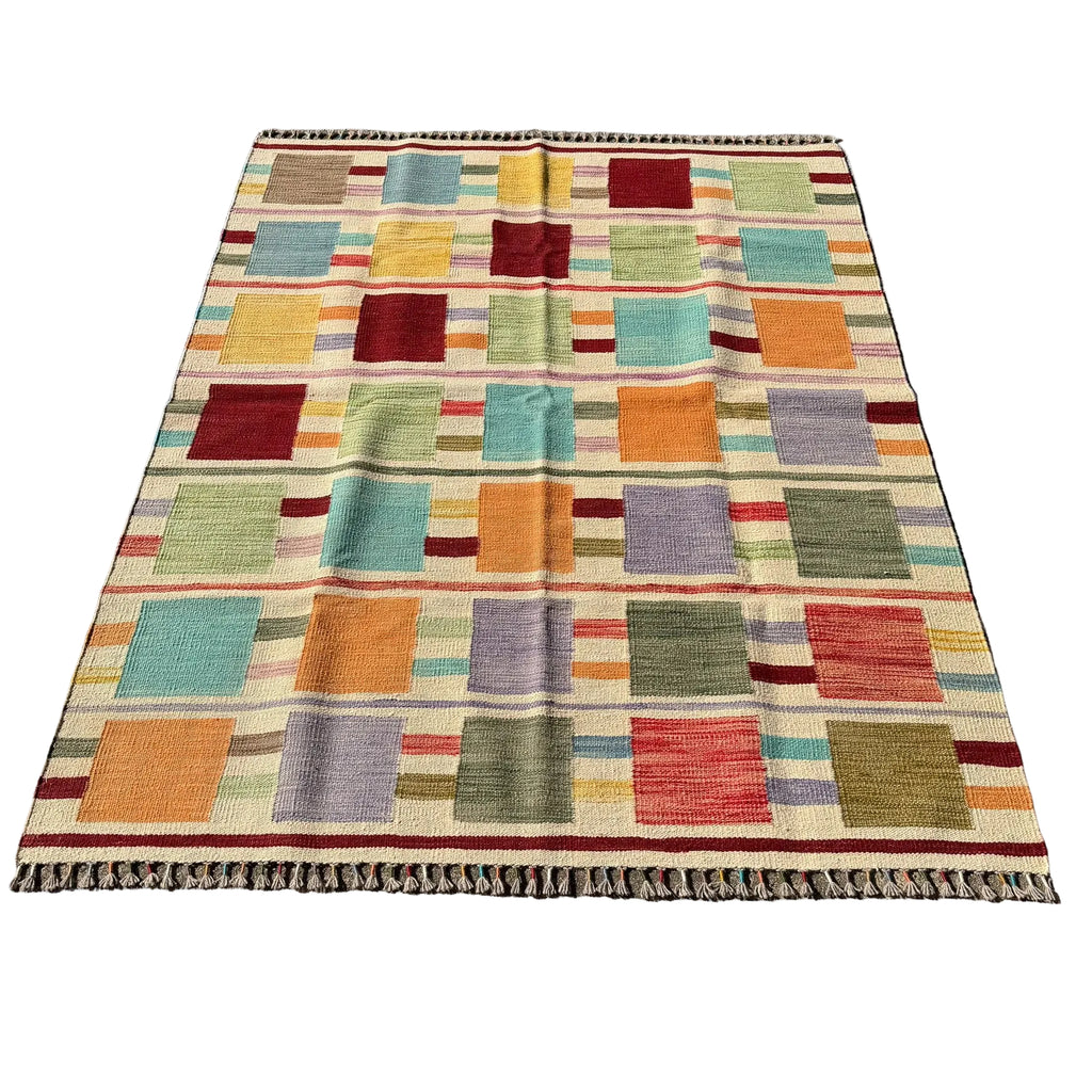 A rectangular woven area rug with a multicolor patchwork design, laid flat on a gray concrete surface. The rug features a grid of colored squares in orange, teal, red, green, purple, and beige, finished with a fringed edge.