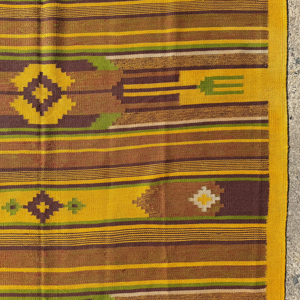 A close-up of a woven textile with horizontal stripes and geometric motifs in yellow, brown, olive green, and cream, photographed against a stone surface.