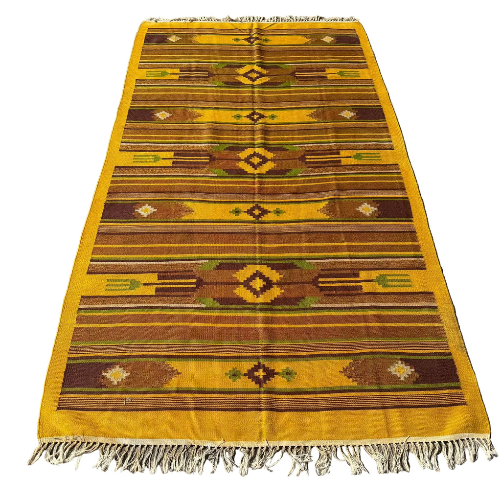 A rectangular woven area rug with fringe, laid flat on a gray concrete surface. The rug features a Southwestern geometric pattern in mustard yellow, brown, olive green, and cream.