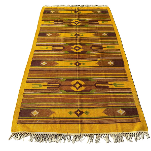 A rectangular woven area rug with fringe, laid flat on a gray concrete surface. The rug features a Southwestern geometric pattern in mustard yellow, brown, olive green, and cream.