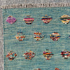 A close-up of a woven rug with a turquoise blue base, featuring rows of small multicolored abstract medallion motifs and a fringed edge.
