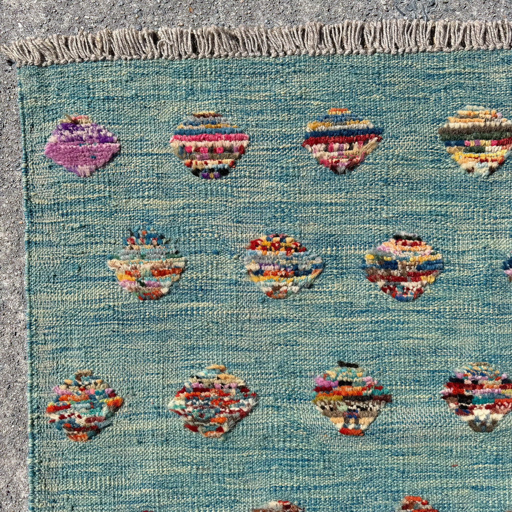 A close-up of a woven rug with a turquoise blue base, featuring rows of small multicolored abstract medallion motifs and a fringed edge.