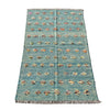 A rectangular woven rug laid flat on a gray concrete surface. The rug has a muted teal-blue base with rows of small multicolored floral or medallion motifs and fringed edges at the top and bottom.