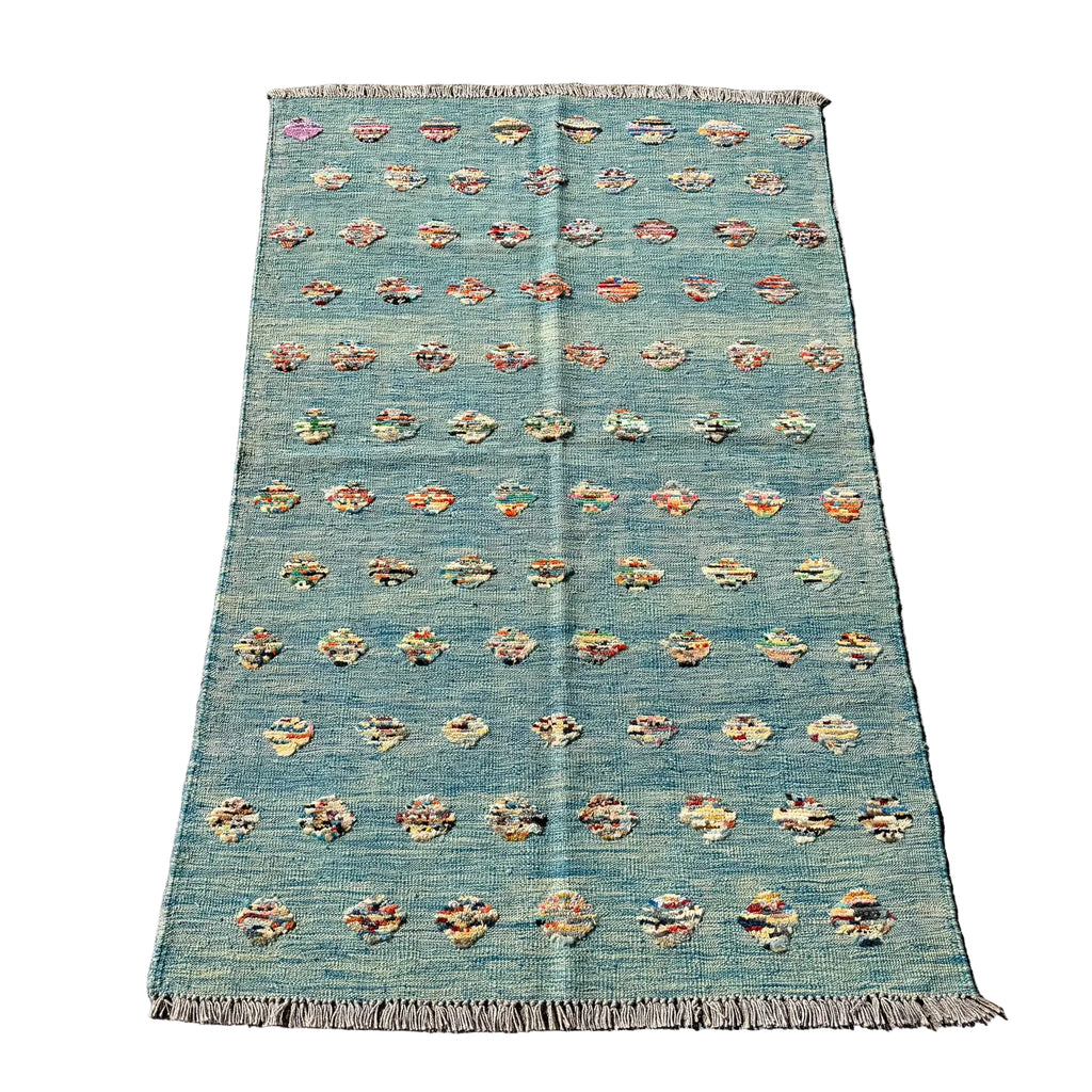 A rectangular woven rug laid flat on a gray concrete surface. The rug has a muted teal-blue base with rows of small multicolored floral or medallion motifs and fringed edges at the top and bottom.