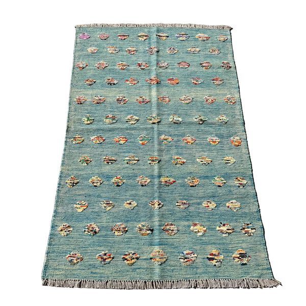 A rectangular woven rug laid flat on a gray concrete surface. The rug has a muted teal-blue base with rows of small multicolored floral or medallion motifs and fringed edges at the top and bottom.