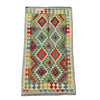 A rectangular kilim rug with a geometric diamond pattern in red, blue, olive green, and beige, laid flat on a textured gray concrete surface.