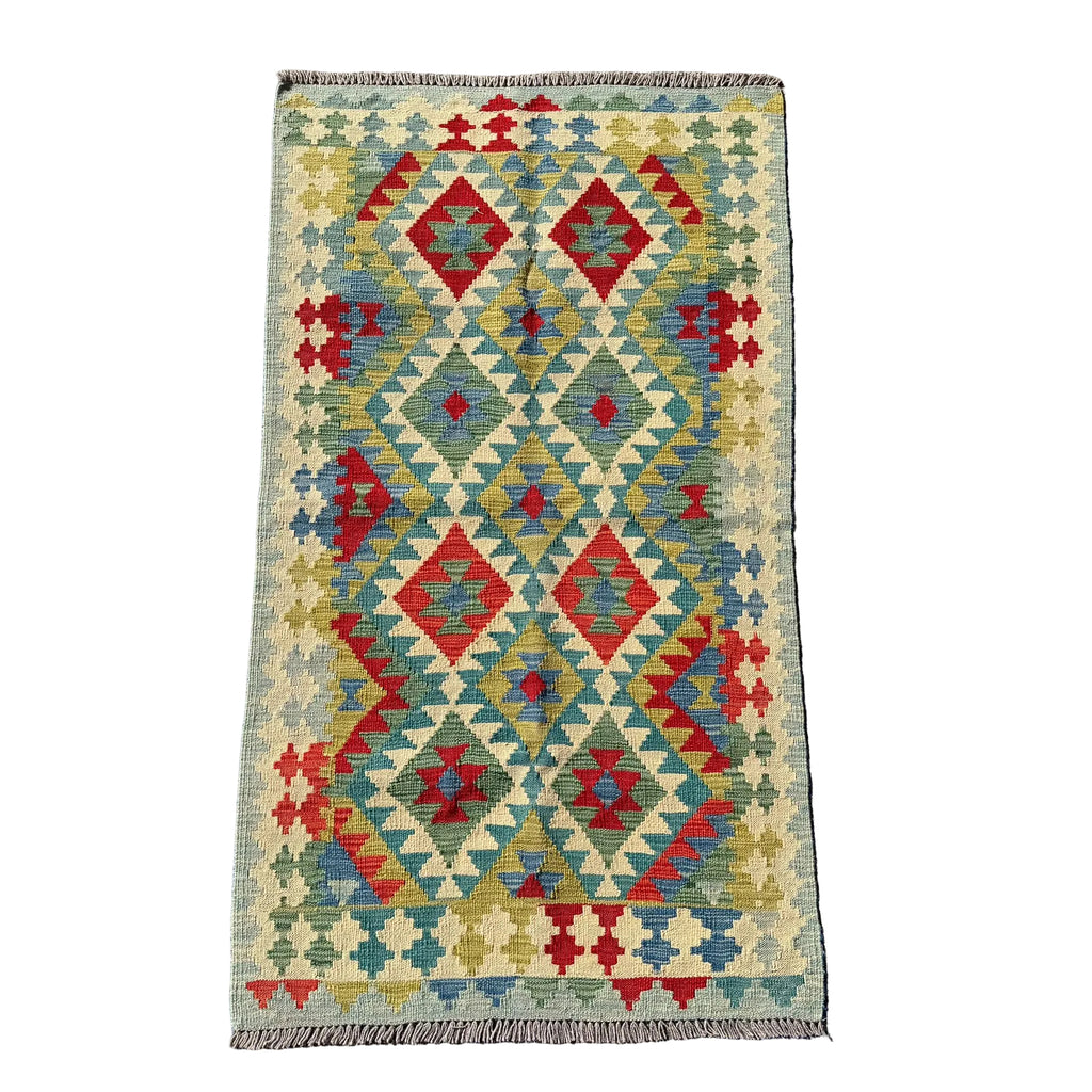 A rectangular kilim rug with a geometric diamond pattern in red, blue, olive green, and beige, laid flat on a textured gray concrete surface.