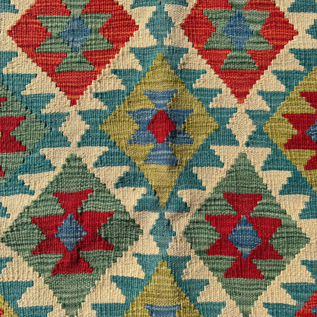 A close-up of a woven textile with a geometric kilim pattern in red, teal, sage green, mustard yellow, and cream.