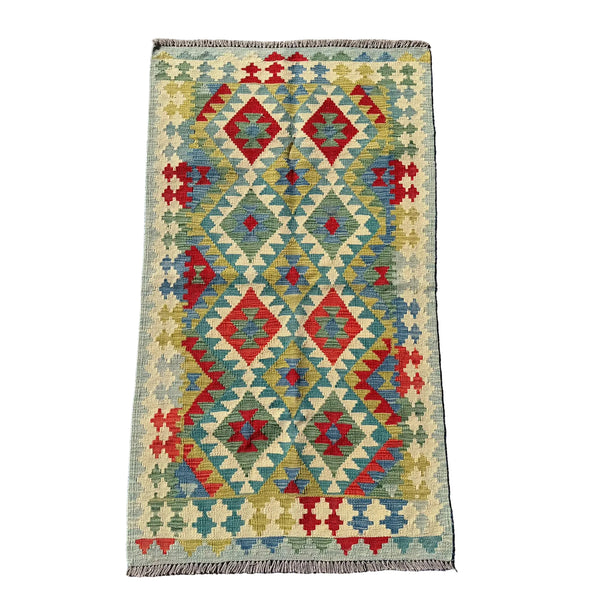 A rectangular kilim rug with a geometric diamond pattern in red, blue, olive green, and beige, laid flat on a textured gray concrete surface.