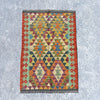 A rectangular kilim rug with a colorful geometric diamond pattern, laid flat on a textured gray concrete surface.