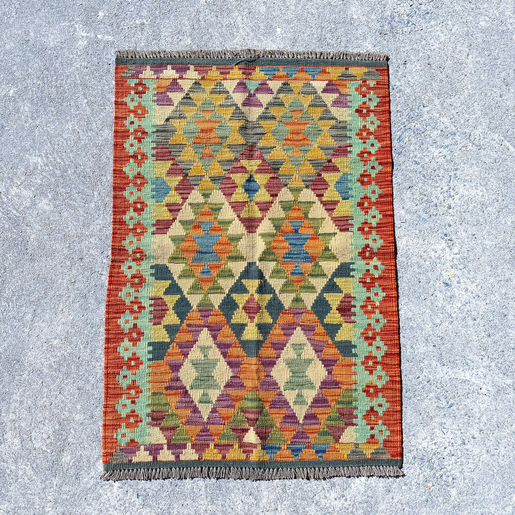 A rectangular kilim rug with a colorful geometric diamond pattern, laid flat on a textured gray concrete surface.
