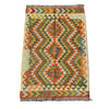 A rectangular woven kilim rug with a geometric diamond pattern in orange, olive green, beige, and blue, laid flat on a gray concrete surface.