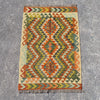 A rectangular woven kilim rug with a geometric diamond pattern in orange, olive green, beige, and blue, laid flat on a gray concrete surface.