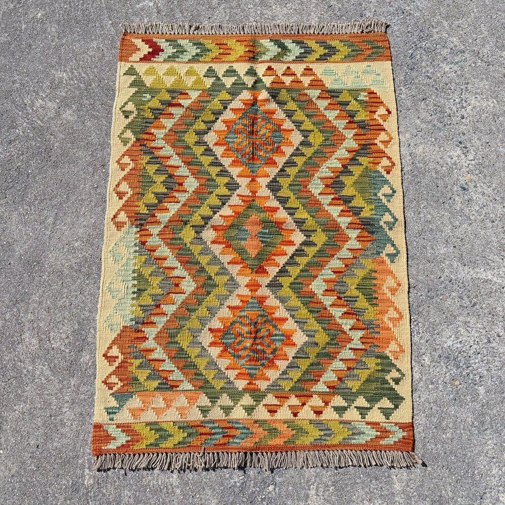 A rectangular woven kilim rug with a geometric diamond pattern in orange, olive green, beige, and blue, laid flat on a gray concrete surface.