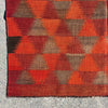 A close-up of a woven kilim rug with a geometric pattern of red, orange, brown, and beige triangles, laid flat on a gray concrete surface.