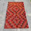 A rectangular woven kilim rug with a geometric triangle pattern in red, orange, and brown, laid flat on a gray concrete surface.