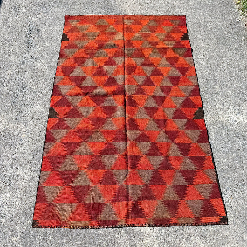 A rectangular woven kilim rug with a geometric triangle pattern in red, orange, and brown, laid flat on a gray concrete surface.