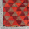 A close-up of a woven textile with a repeating geometric triangle pattern in red, rust orange, and taupe-brown, laid flat on a gray concrete surface.