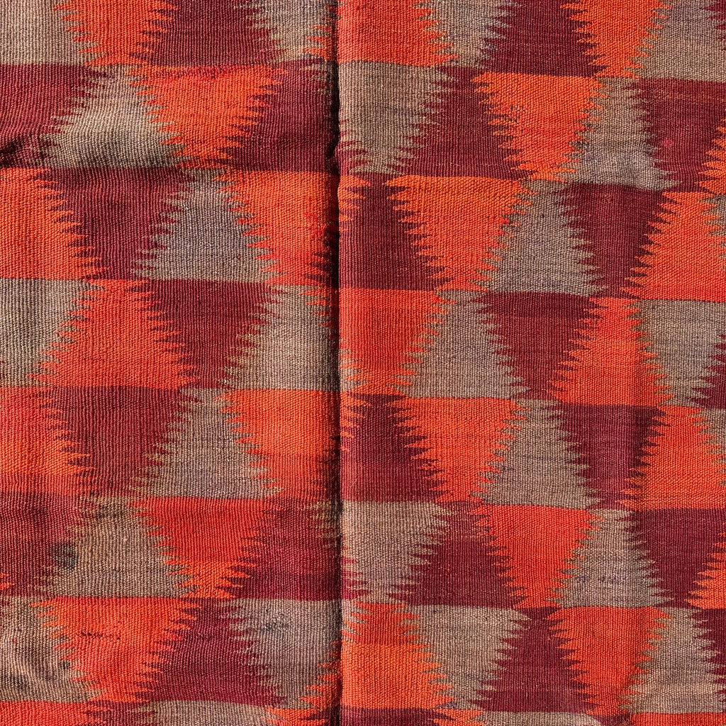 A close-up of a woven textile with a repeating geometric pattern of orange, rust red, and taupe-brown triangles.