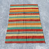 A rectangular striped wool rug laid flat on a gray concrete surface. The rug features horizontal bands of rust red, teal green, orange, and natural beige with a fringed edge.