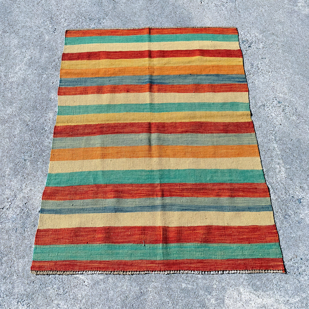 A rectangular striped wool rug laid flat on a gray concrete surface. The rug features horizontal bands of rust red, teal green, orange, and natural beige with a fringed edge.