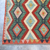 A close-up of a woven kilim rug showing its geometric diamond pattern in red, green, cream, light blue, and orange, with a fringed edge on a concrete floor.